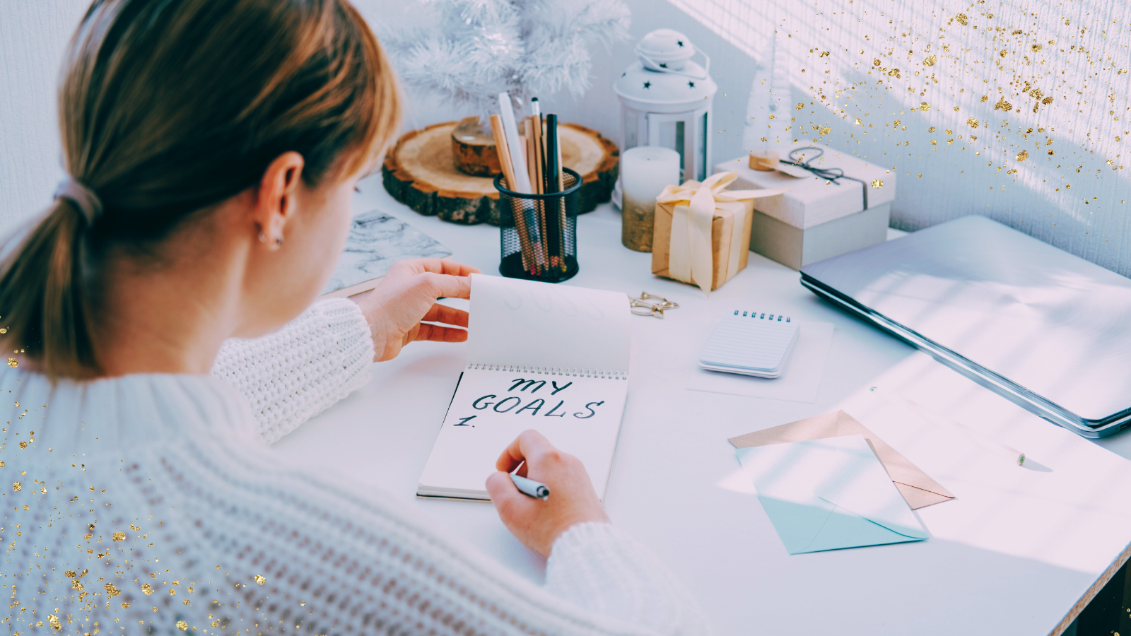 A woman sitting at a desk writing in a notebook labeled "My Goals" in bold letters. The desk is neatly organized with a small tree decoration, a container of pens, wrapped gifts, and a laptop. Soft natural light illuminates the scene, with golden glitter accents adding a festive or motivational touch.