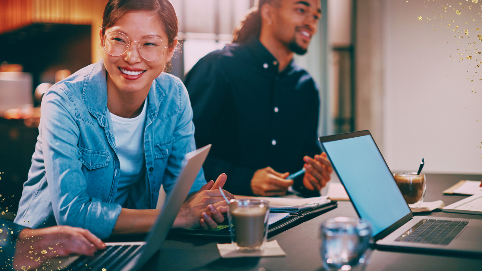 A cheerful team meeting in a modern workspace. A smiling woman wearing glasses and a denim shirt leans forward while engaging with colleagues. Two open laptops, coffee cups, and a notebook are on the table, creating a collaborative and relaxed atmosphere. A man in the background is holding a pen and also smiling, contributing to the positive energy of the discussion. Golden glitter accents add a celebratory or motivational vibe to the scene.