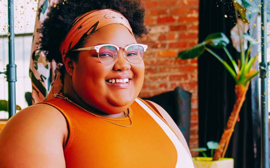 A cheerful woman wearing glasses, a patterned headscarf, and an orange sleeveless top smiles warmly in a cozy, modern workspace. The background features an exposed brick wall, large windows, and vibrant green plants, creating a welcoming and creative atmosphere. Golden glitter accents add a festive or inspirational touch to the image.