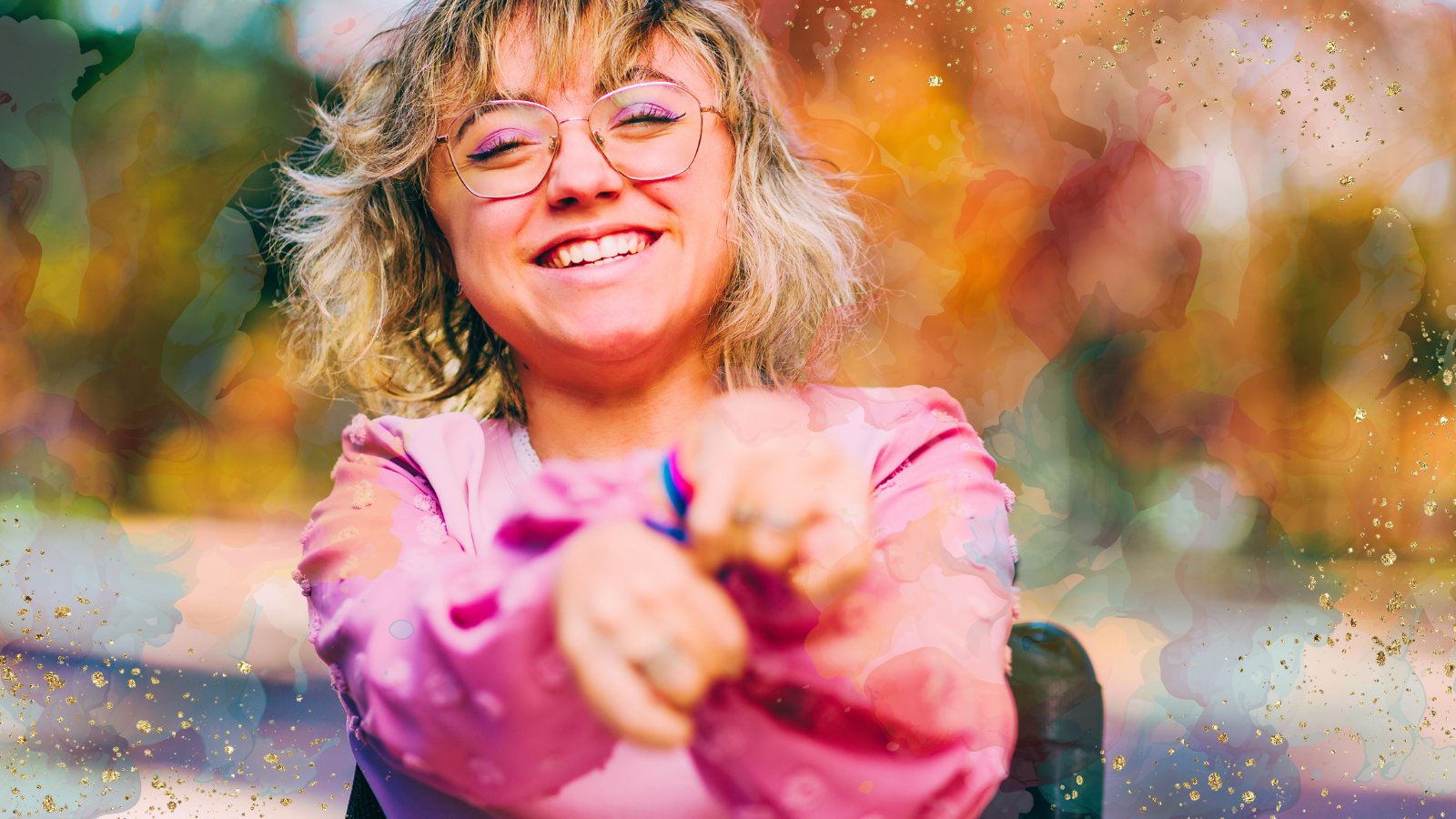 A cheerful woman wearing glasses and a pink blouse smiling brightly outdoors. She has her arms extended playfully towards the camera. The background features blurred autumn colors with vibrant watercolor splashes and golden accents, adding a creative and joyful feel to the image.