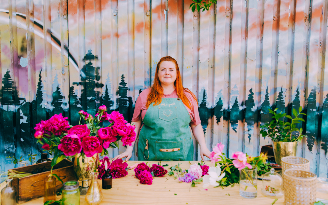 A woman with long red hair stands confidently behind a wooden table filled with vibrant pink and white flowers, greenery, and glass vases. She wears a green apron over a pink shirt, with a painted mural of a sunset and trees on a corrugated metal wall in the background.