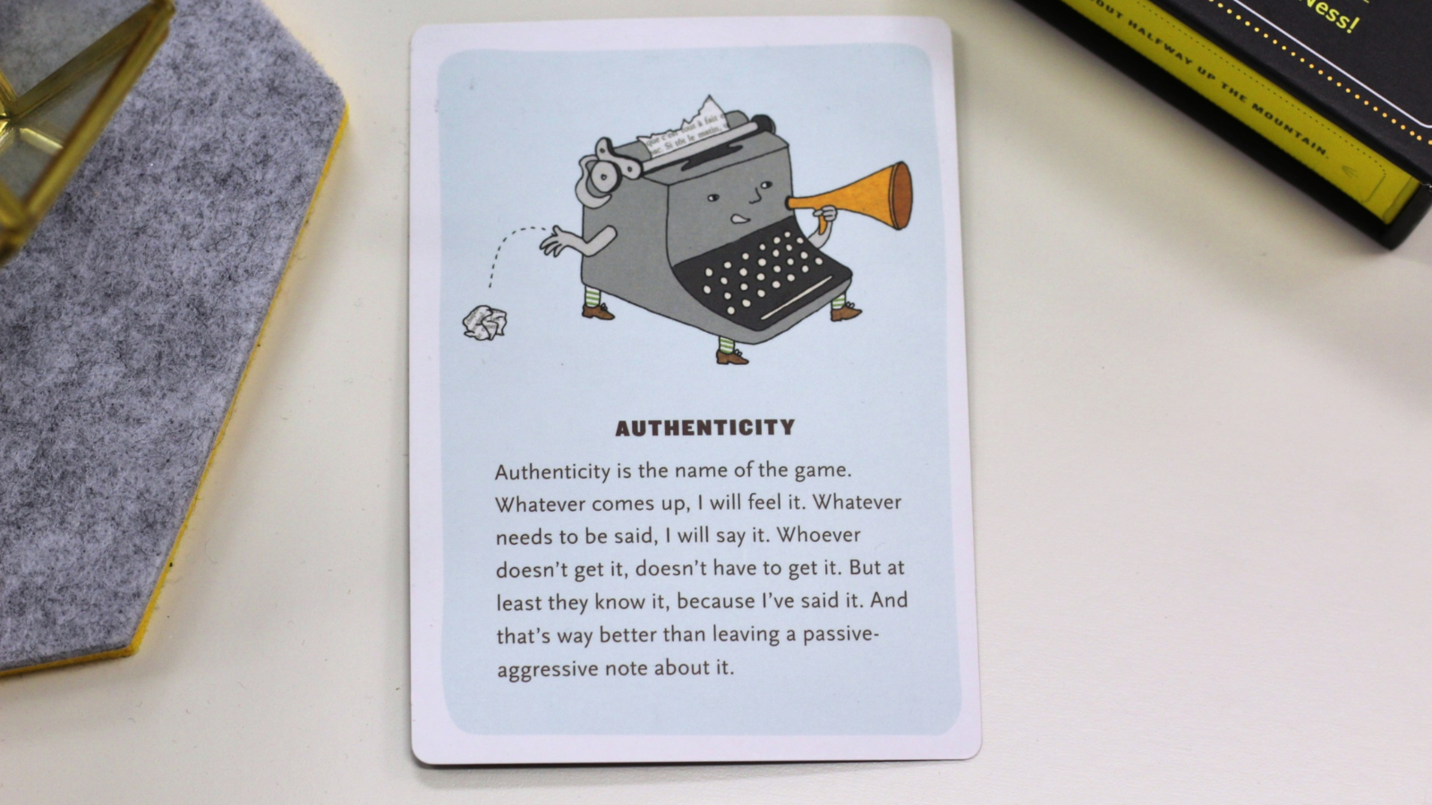 A card from the 'Affirmators!' affirmation deck is placed on a white surface. The card is titled 'Authenticity' and features a whimsical illustration of an anthropomorphized vintage typewriter with arms, legs, and a face. The typewriter is holding a megaphone in one hand and tossing a crumpled piece of paper with the other. The background of the card is light blue. Below the illustration, the text reads: 'Authenticity is the name of the game. Whatever comes up, I will feel it. Whatever needs to be said, I will say it. Whoever doesn’t get it, doesn’t have to get it. But at least they know it, because I’ve said it. And that’s way better than leaving a passive-aggressive note about it.' The card is placed near a felt coaster and part of the deck's box is visible in the background