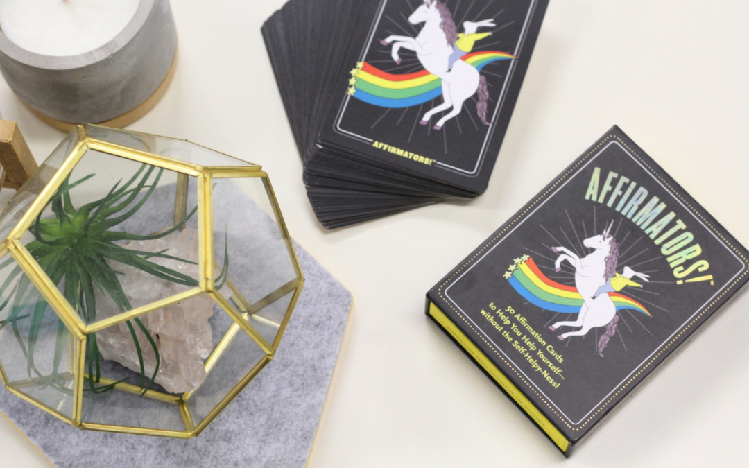 A deck of 'Affirmators!' affirmation cards is displayed on a white surface. The box and cards feature a whimsical design with a white unicorn rearing on its hind legs, a rainbow tail, and yellow stars. The box reads: '50 Affirmation Cards to Help You Help Yourself—Without the Self-Helpy-Ness!' Nearby, a geometric glass terrarium with gold edges contains a green air plant and a rose quartz crystal. A concrete candle holder and a felt coaster are also present, creating a calming and aesthetically pleasing arrangement.