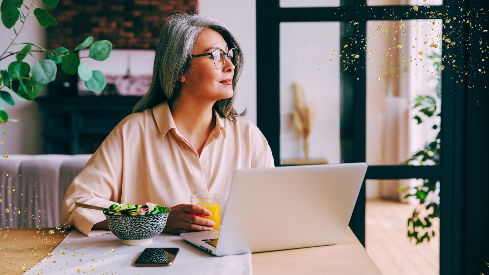 A woman with long, silver hair and glasses sits at a table, gazing thoughtfully out of a window. She is wearing a light blouse and has a laptop open in front of her, along with a bowl of salad, a glass of orange juice, and a smartphone resting nearby. The background features soft natural light, greenery, and cozy decor, with golden sparkles subtly overlaying the image.