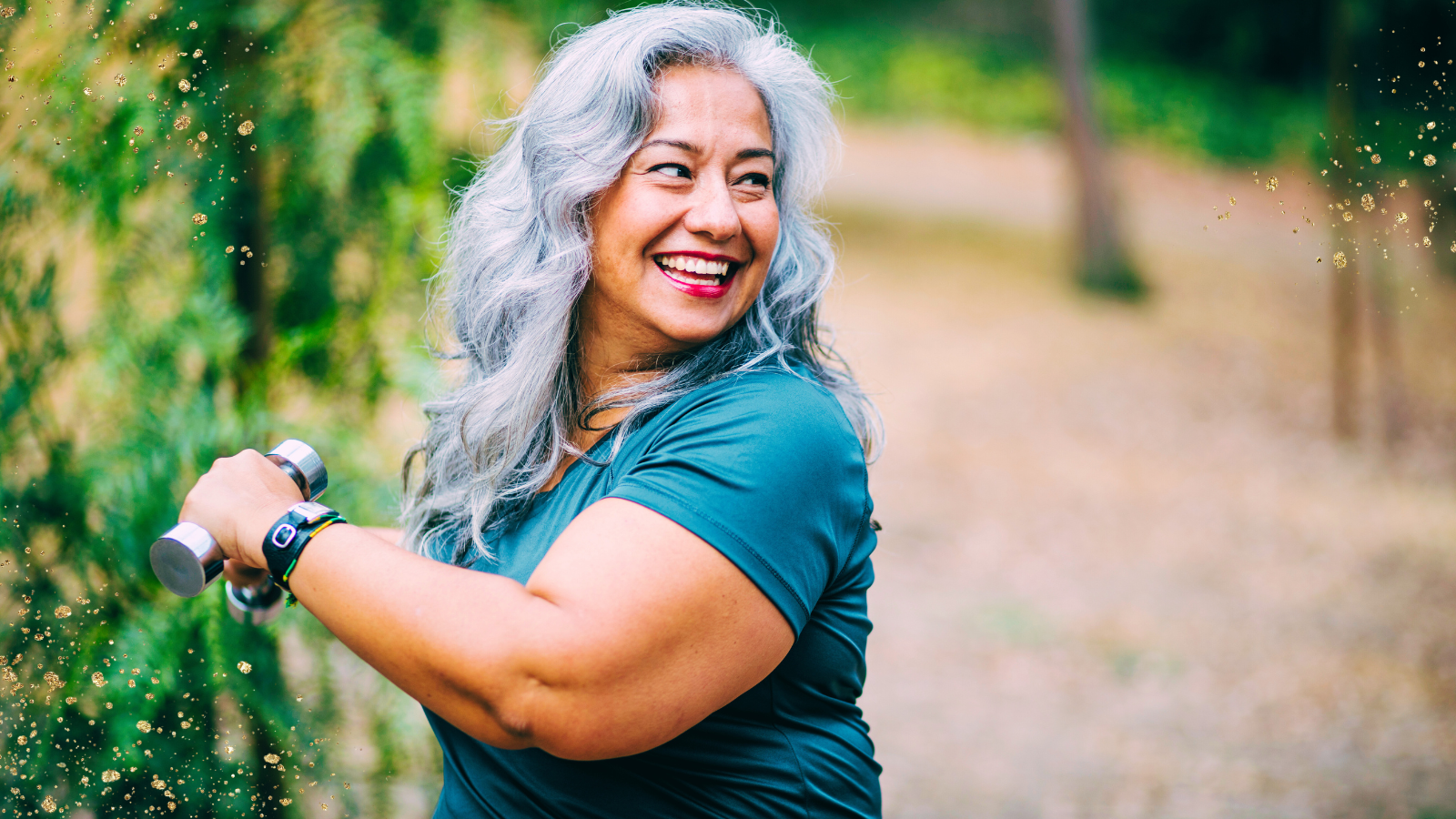 A woman with long gray hair smiles brightly while exercising outdoors. She is holding a pair of dumbbells and wearing a teal athletic top and a fitness tracker on her wrist. The background features a blurred, natural setting with greenery and trees, creating a vibrant and energetic atmosphere. Subtle gold sparkles are sprinkled across the image, adding a touch of creativity.