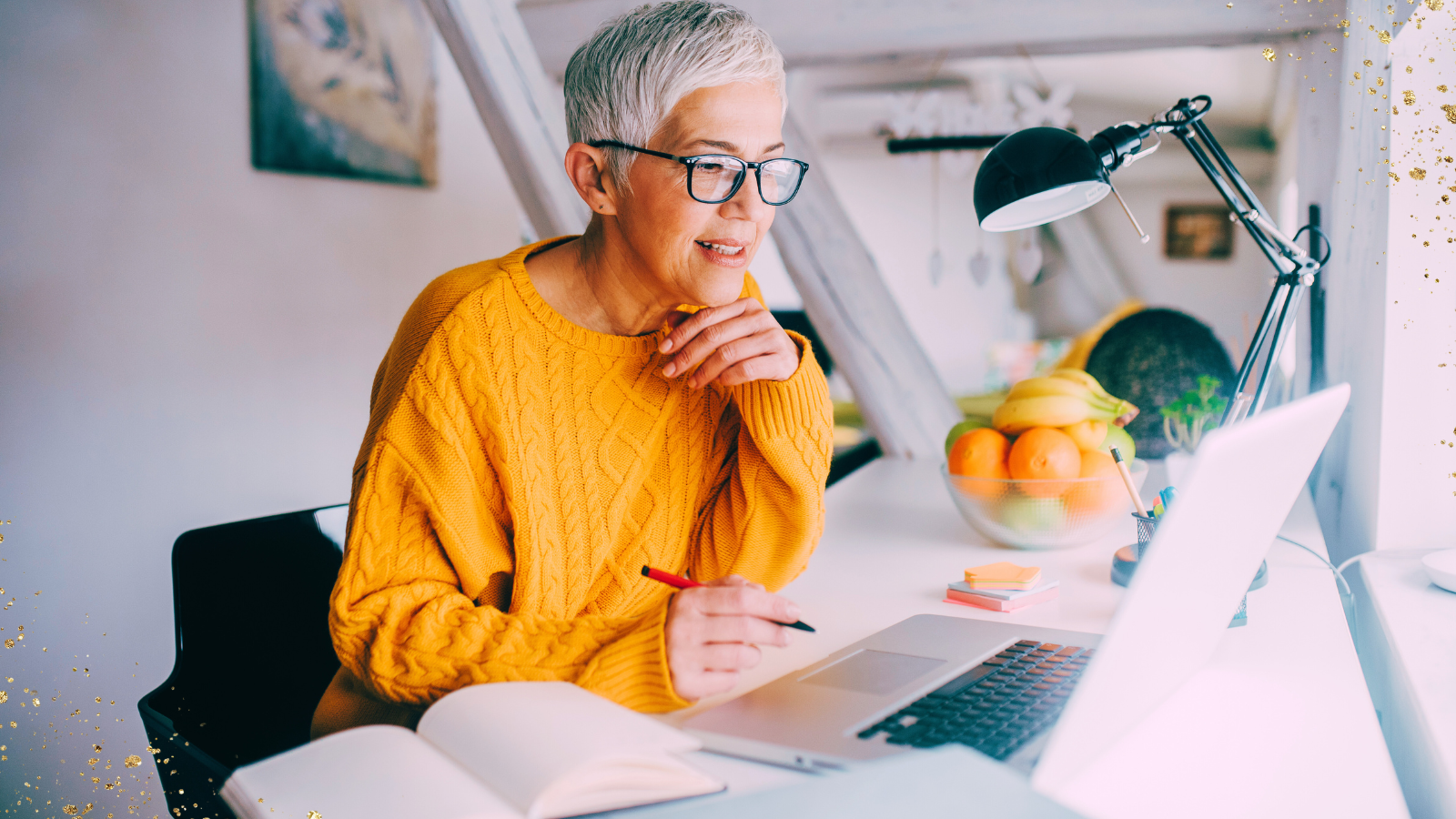 A woman with short white hair and glasses is sitting at a bright desk, smiling as she looks at her laptop. She is wearing a cozy yellow sweater and holding a pen, with an open notebook in front of her. A modern desk lamp, a bowl of fresh fruit, and a few colorful sticky notes are neatly arranged on the desk. The background features a softly lit room with light wooden beams and a warm, inviting atmosphere. Gold sparkles add a creative accent to the image.