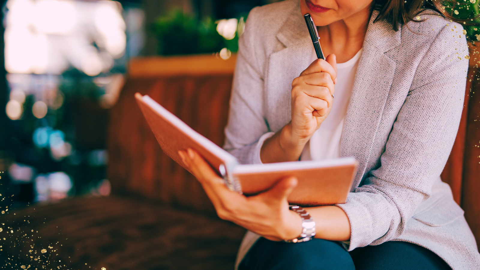 Close-up of a woman sitting on a cushioned bench, holding a pen to her lips in thought while reading or writing in a notebook. She is dressed in a light blazer and dark pants, with a watch on her wrist. The background is softly blurred with warm lighting and greenery, adding a cozy and focused atmosphere, complemented by golden sparkles lightly overlaying the image.