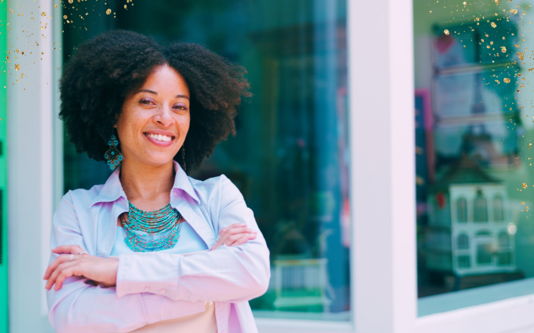 A confident woman with natural curly hair stands smiling outside a storefront with glass windows. She is wearing a light-colored shirt and a layered turquoise necklace, with her arms crossed. The background includes a glimpse of colorful decorations and a bright teal door, with golden sparkles lightly overlaying the image